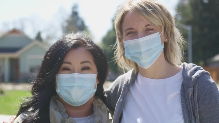 Asian woman with straight black hair and white woman with dirty blonde hair wear light blue masks and face the camera with eye contact