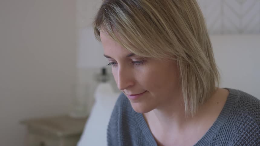 Blonde woman with a grey sweater sits on a bed and uses a laptop in the bedroom