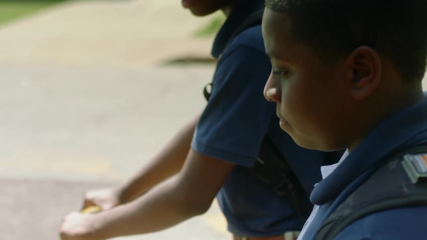 A pair of boys wearing backpacks walk along a sidewalk through a neighborhood while one pushes a bicycle