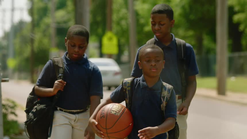 A group of boys in school uniforms walk along a sidewalk through a neighborhood during the day