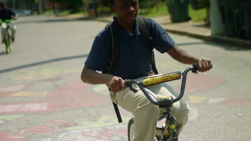 A pair of boys wearing backpacks ride down a street on bicycles on a sunny day