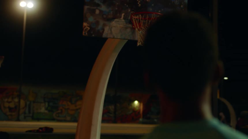 A teenage boy at an outdoor basketball court looks up at the basketball net