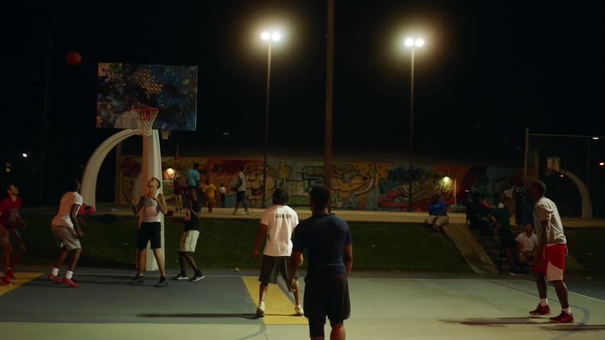 Men play basketball on park court at night as others watch