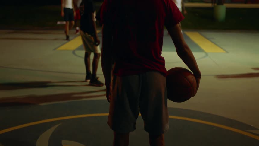 A basketball player stands on the sidelines with a ball under his right arm as people play the game