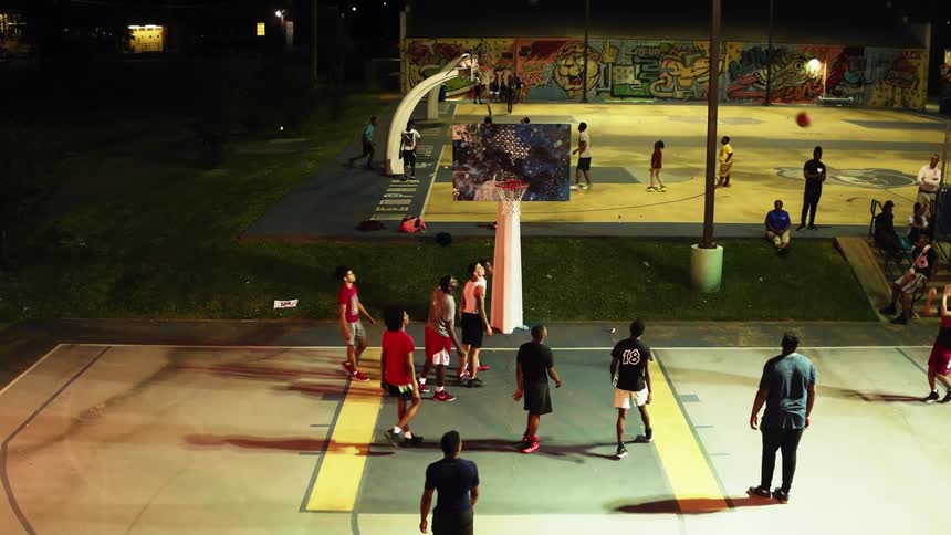 Teenagers play basketball on two outdoor courts as a few people watch