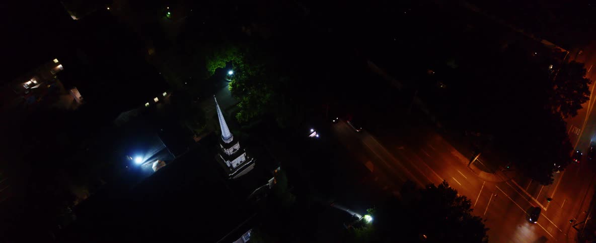 Cars drive along the streets of a downtown area at night while near a building with a tall steeple