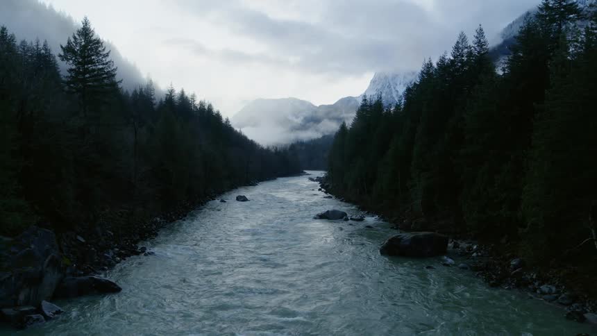 A fast flowing river surrounded by trees with a mountain range, mist and clouds in the background