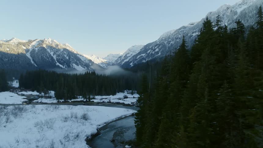 Revealing a snow covered mountain range and river valley from behind a stand of fir trees