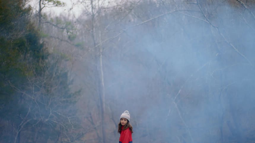A young girl stands in a forest and turns around as smoke rises around her