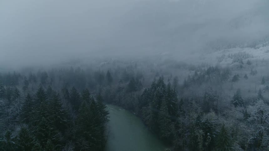 Aerial view of a river flowing through a misty mountain forest while it snows lightly