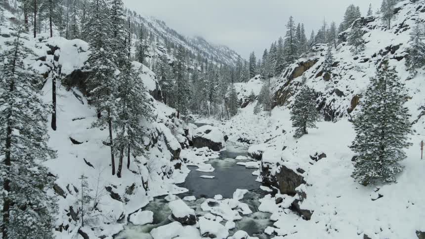 Flying upstream above a beautiful mountain valley river partially frozen and dotted with snow