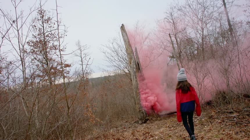 Little girl wearing a jacket and beanie walks towards bright pink smoke coming out of a building in the forest