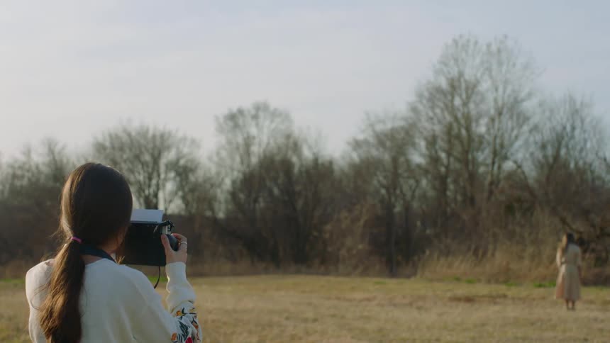 A woman with a polaroid camera taking a picture of another woman with trees and the sky in the background