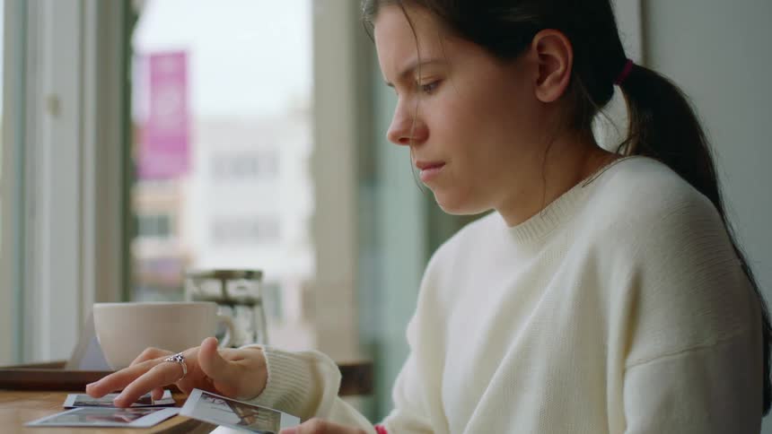 Young woman wearing a white sweater checks polaroid pictures on the table of a restaurant
