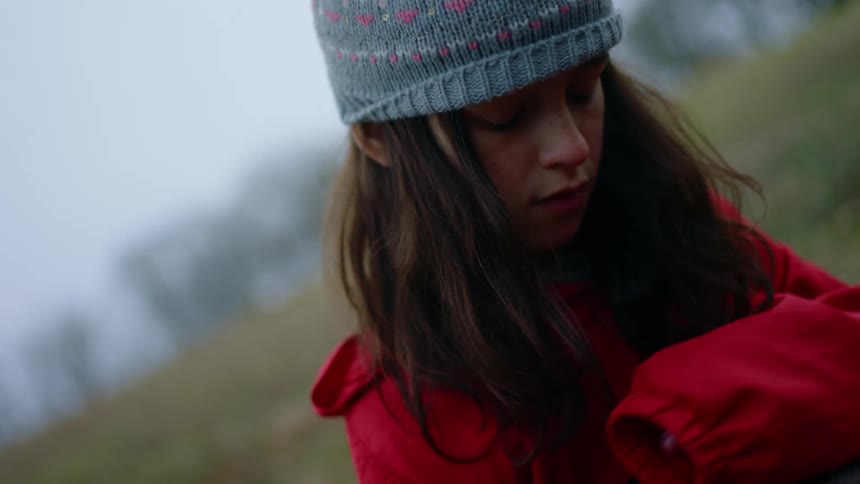 Girl wearing a red oversized jacket and blue beanie pets a cat in the grass