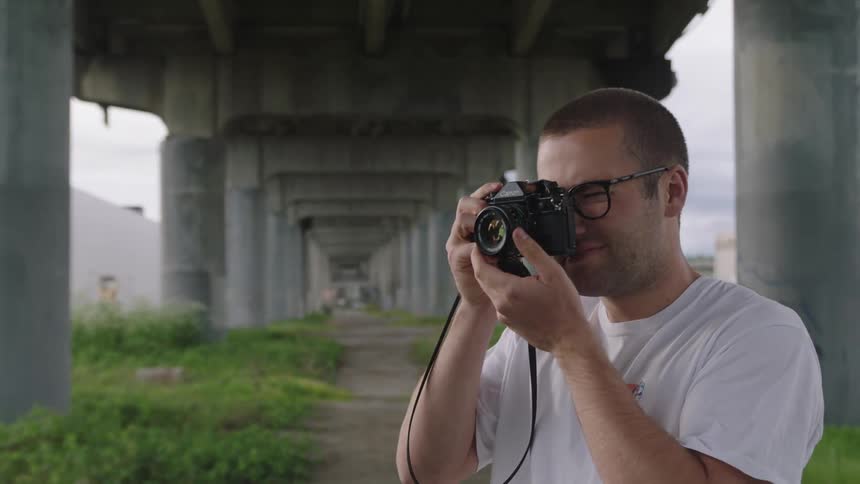 A man holds a camera up to his face standing under a rural bridge
