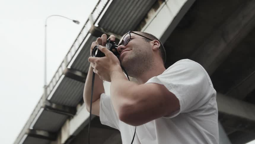 A male photographer standing under a bridge takes a photo with a camera before lowering it from his face