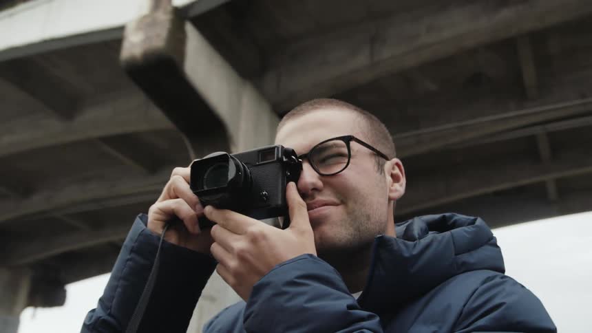 A male photographer takes a photo with a camera while standing under a concrete bridge