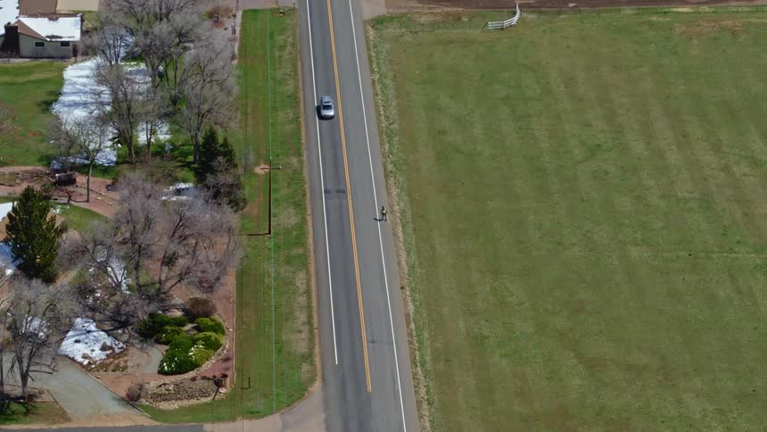 Two cars pass each other on a rural road near farm fields and homes