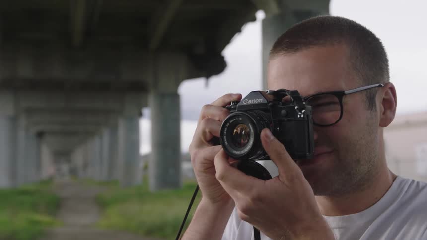 Young man with glasses and scruffy beard is under a bridge taking photos of something out of sight on the left
