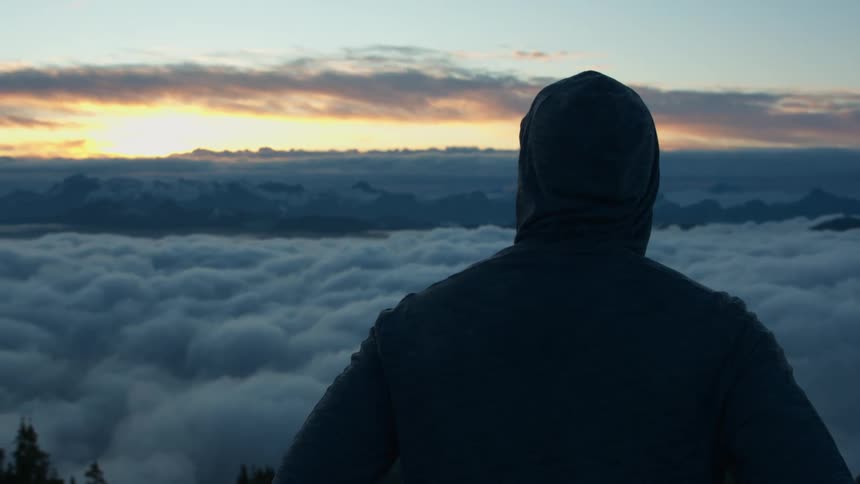 A person in a hoodie stands on a mountain and looks out at the sea of clouds obscuring the landscape below