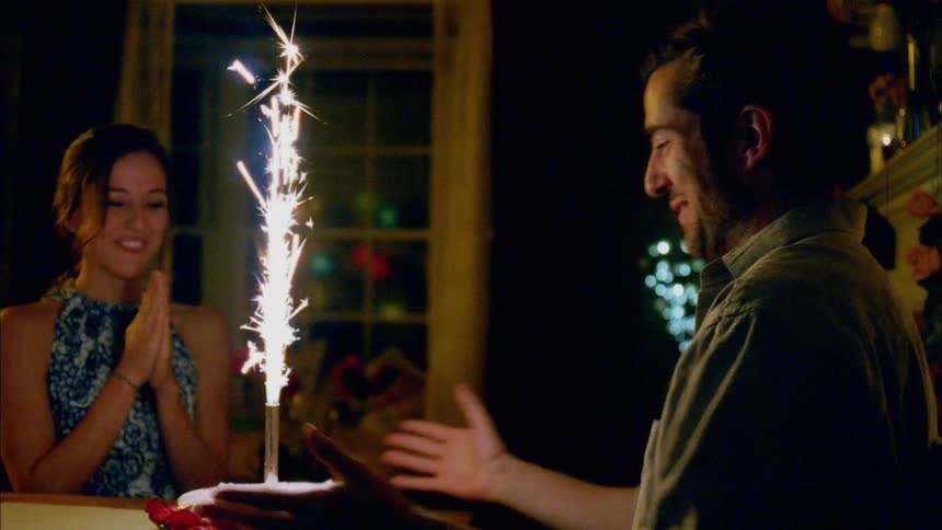 A woman brings a man a cake with a large flare on top and he attempts to it out as they laugh