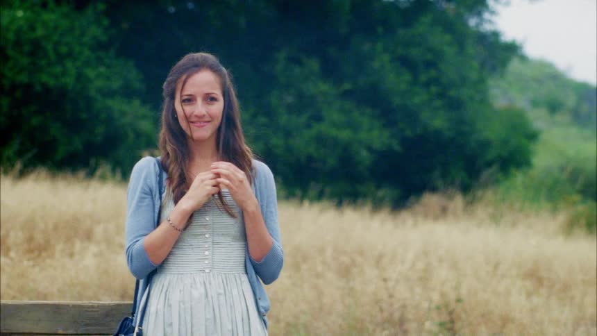 A young woman stands against a wooden fence in a field of yellow grass and smiles as she fiddles with her hair