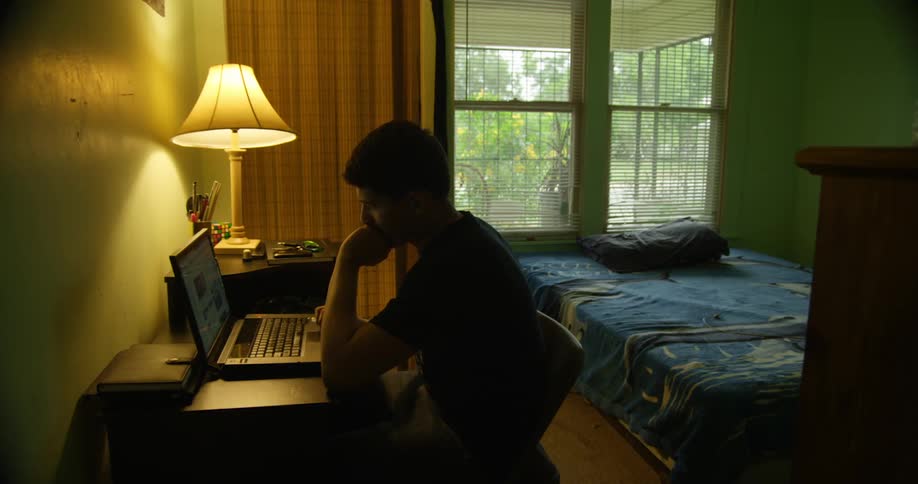 Man with a black t shirt sits at a desk and uses a laptop in his room