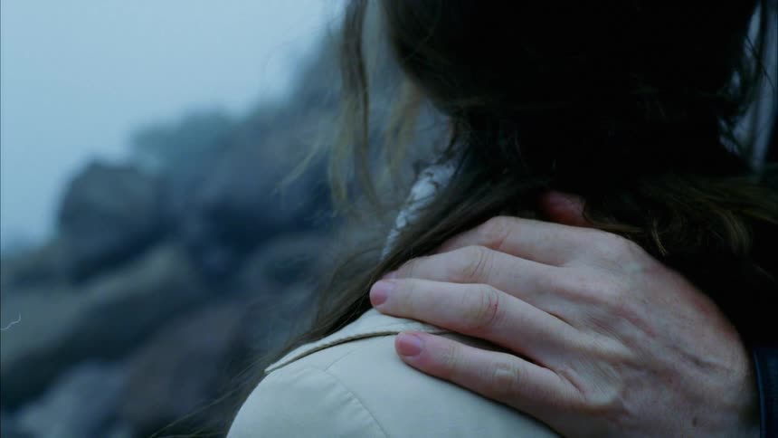 A man puts his hand on his wifes shoulder as they stand on rocks overlooking the shore