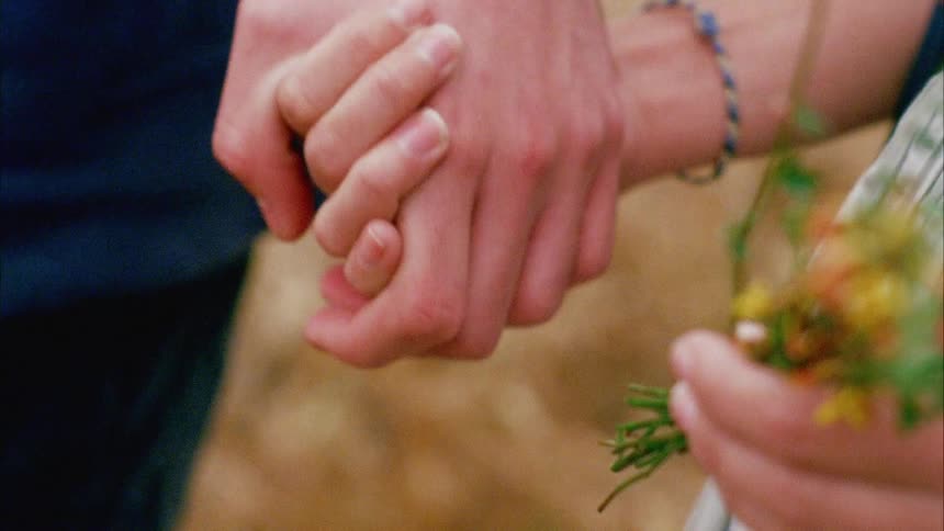 A woman carrying wildflowers walks with a man who reaches over and takes her hand