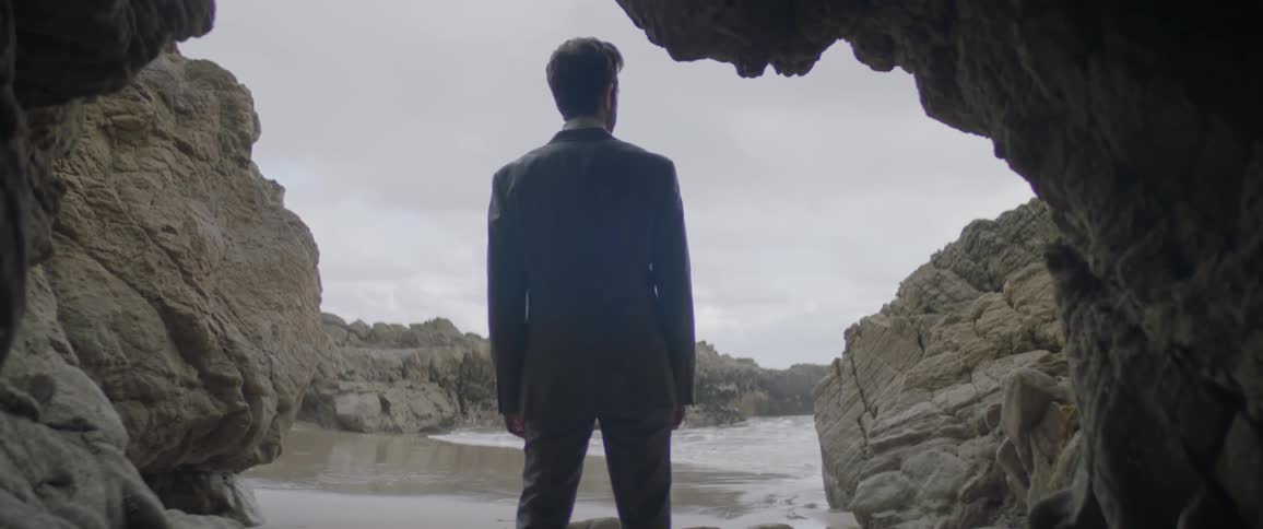 Man stands in the mouth of a cave looking out at a body of water