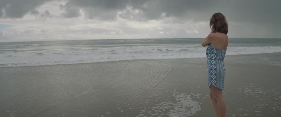 Woman in dress standing on beach while waves wash up on cloudy day