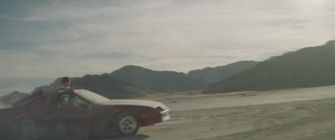 A man is clinging to the top of a car as it speeds in circles in the desert during the day