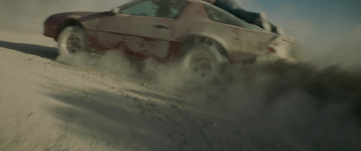 Woman driving erratically in a dusty area and enjoying it while a man clings to the top of the car near sunset