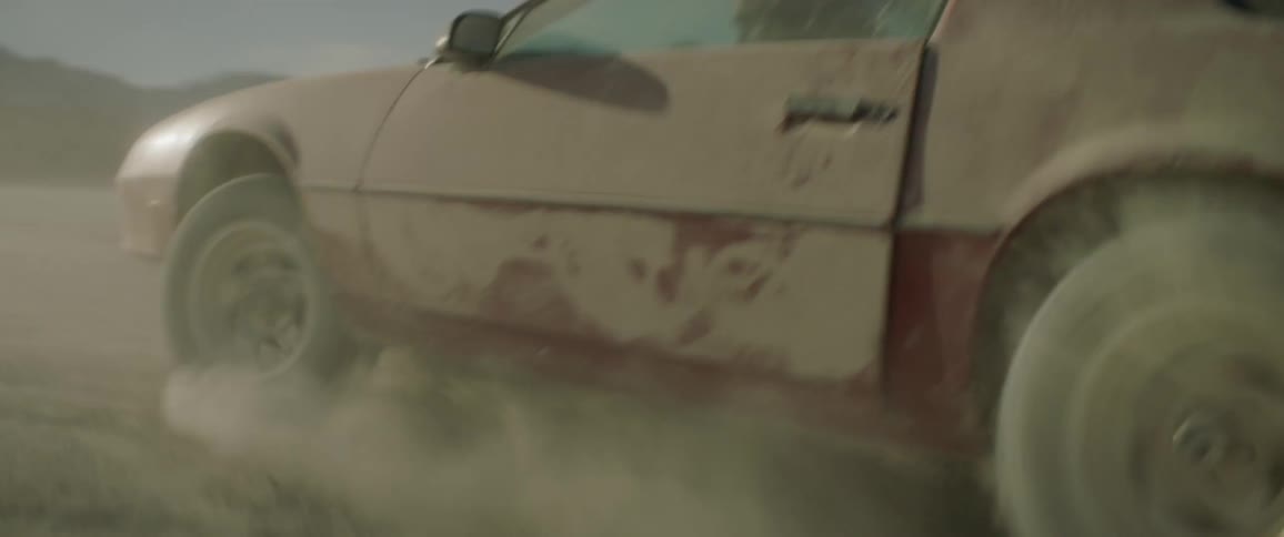 Woman driving erratically in a dusty area and enjoying it while a man clings to the top of the car near sunset