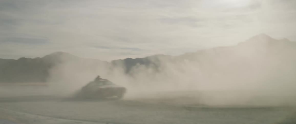 A red car does donuts in the dirt as a man holds onto the roof