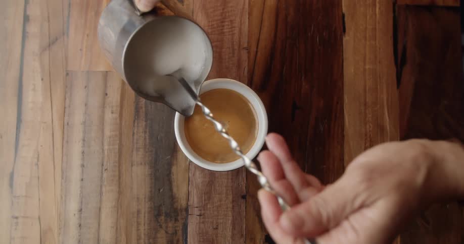 A barista adds steamed milk to a cup of a coffee on a wood counter