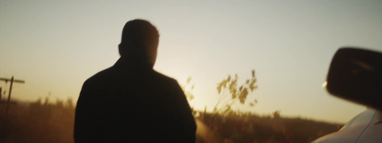 A man climbs out of a truck parked in front of a hill and walks around the front of the vehicle as the sun shines over the hill