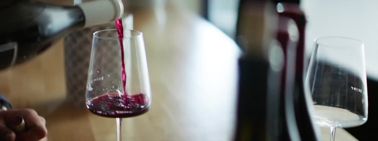 A person pours red wine from a wine bottle into a wine glass resting on a wooden counter