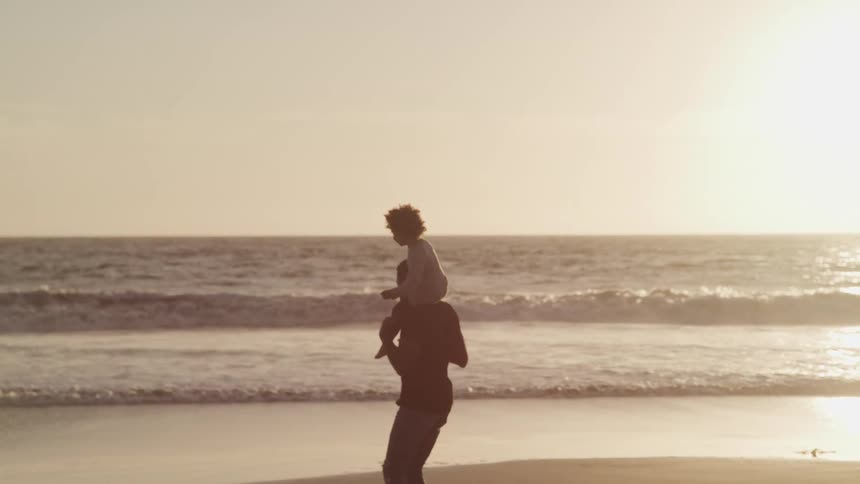 A man spins while carrying a child on his shoulder on the beach as the sun shines over the horizon