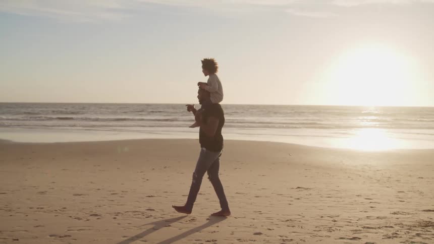 Father gives son a piggyback ride as they walk down the beach at sunrise and point to something in the distance