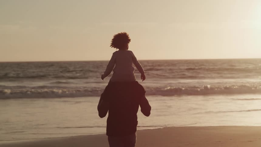 Loving dad carries son in his shoulders while walking on the beach at sunset