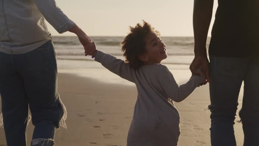 A toddler holds hands with her parents as they walk down the beach