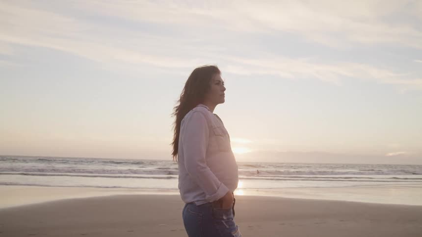 Side view of a woman with a grey shirt and jeans standing on beach at sunset next to the sea