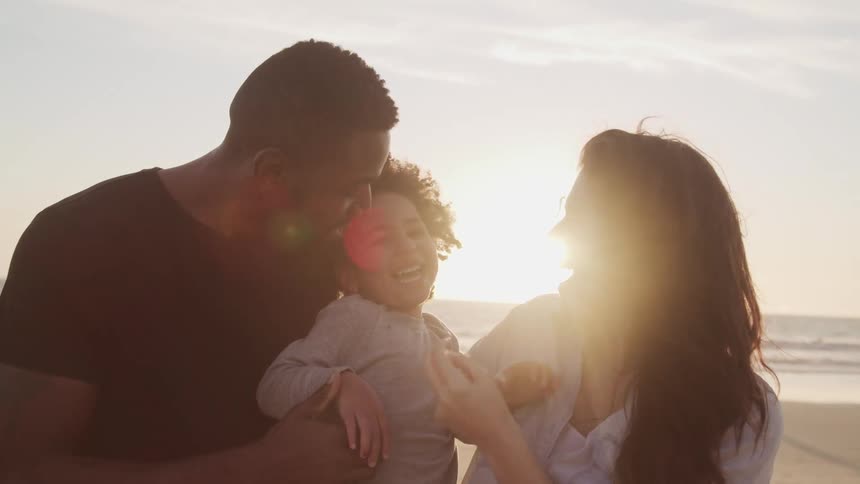 A man holds his young daughter while they and his wife stand on the beach laughing together