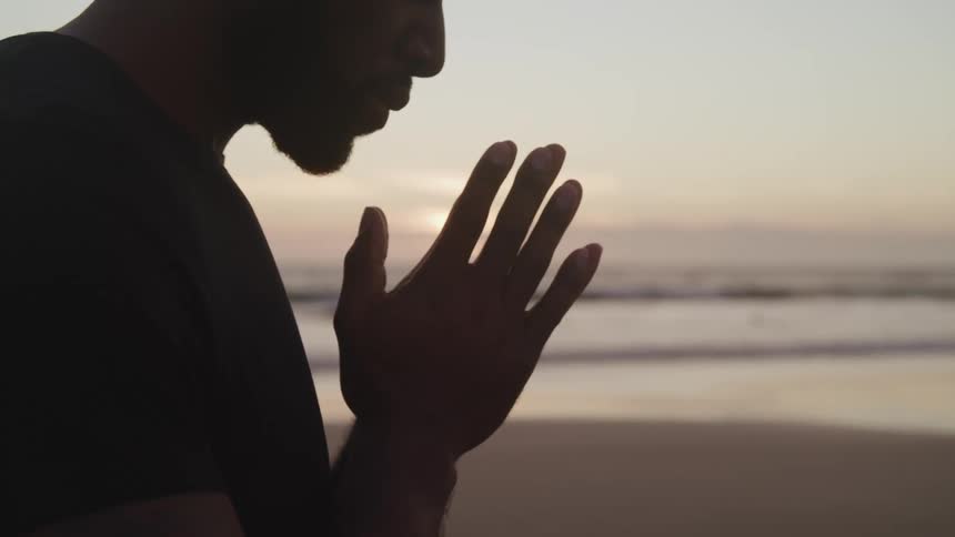 A black man prays while standing on the beach at sunset