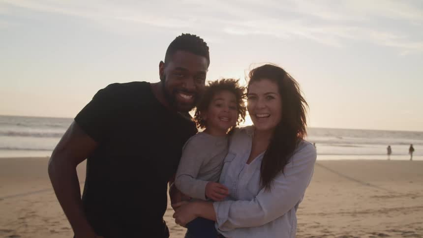 Black father son and white mother pose for the camera on the sand beach at sunset next to the ocean