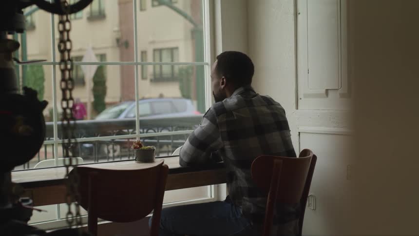 A man sits at a window in a cafe and drinks a coffee as he watches people walk by