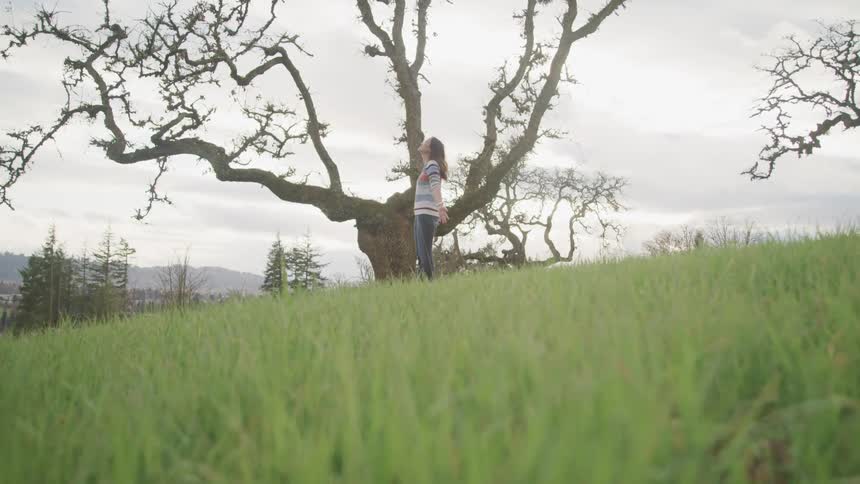 A woman standing in a grassy field with trees the sky and clouds in the background