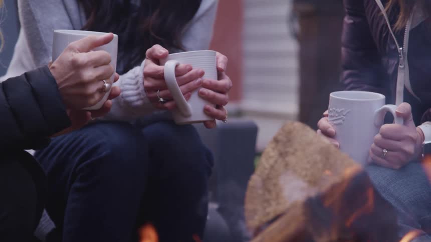 Three women sitting around a fire in a back yard drinking out of mugs
